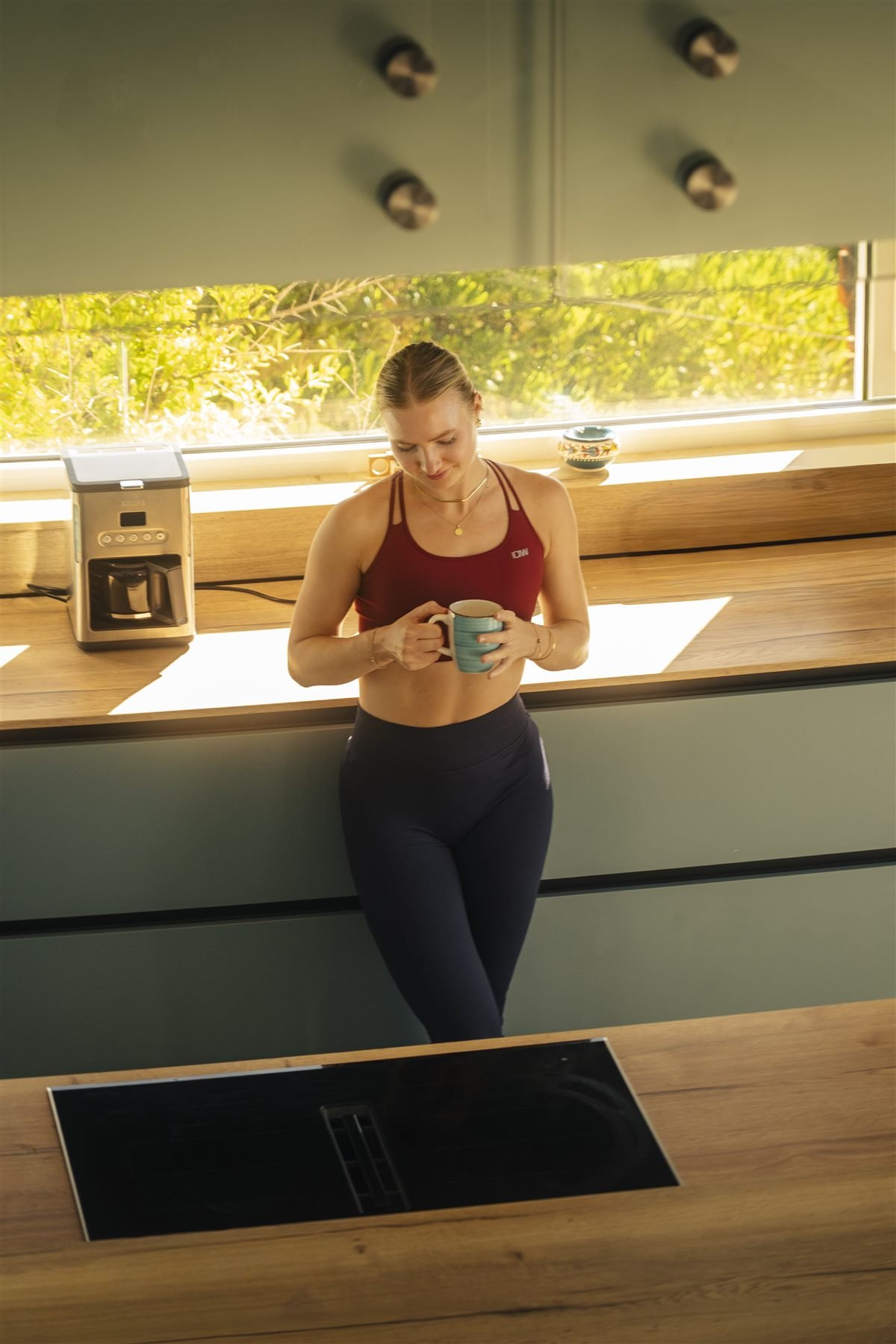 Lilli Schmid lehnt in einer sonnigen Küche an der Arbeitsplatte und hält eine Tasse in den Händen